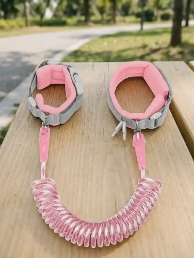 Pink and white coiled child anti-lost wristband with dual padded straps resting on a wooden outdoor table.